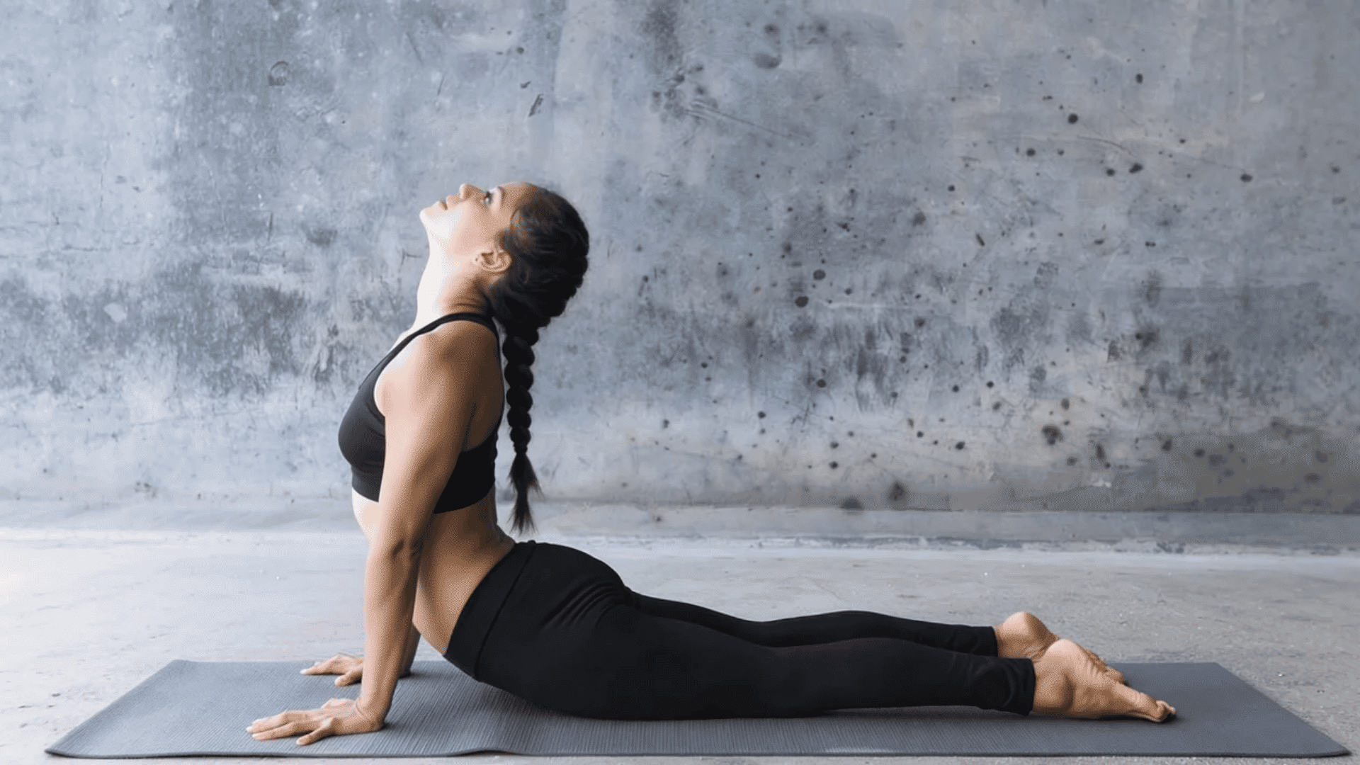Woman performing Cobra Pose (Bhujangasana) on a yoga mat, lifting her chest with straight arms while legs remain extended, set against a textured concrete wall background
