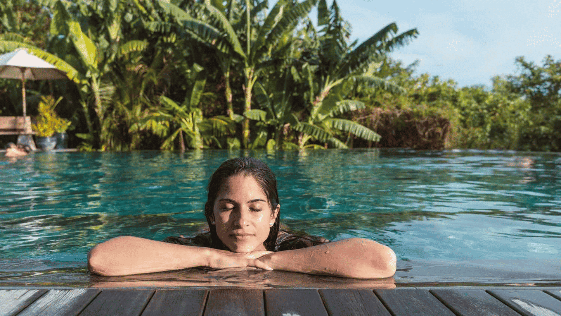 Woman relaxing in an outdoor pool, resting her arms on the edge with eyes closed, surrounded by lush tropical greenery.