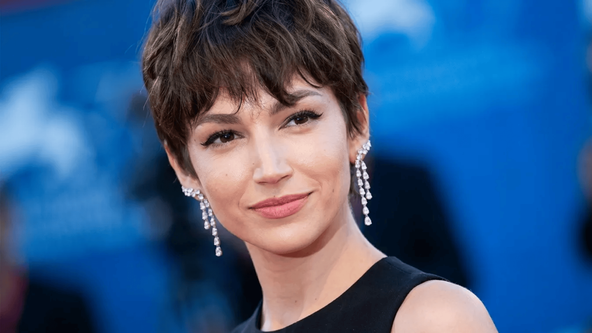 Woman with a short pixie haircut in dark brown, wearing elegant dangling earrings and a black outfit, looking slightly to the side against a blue event-style background.