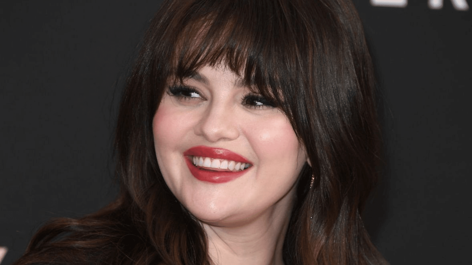Woman with long dark hair and soft, wispy bangs, styled in loose waves with a side-swept finish