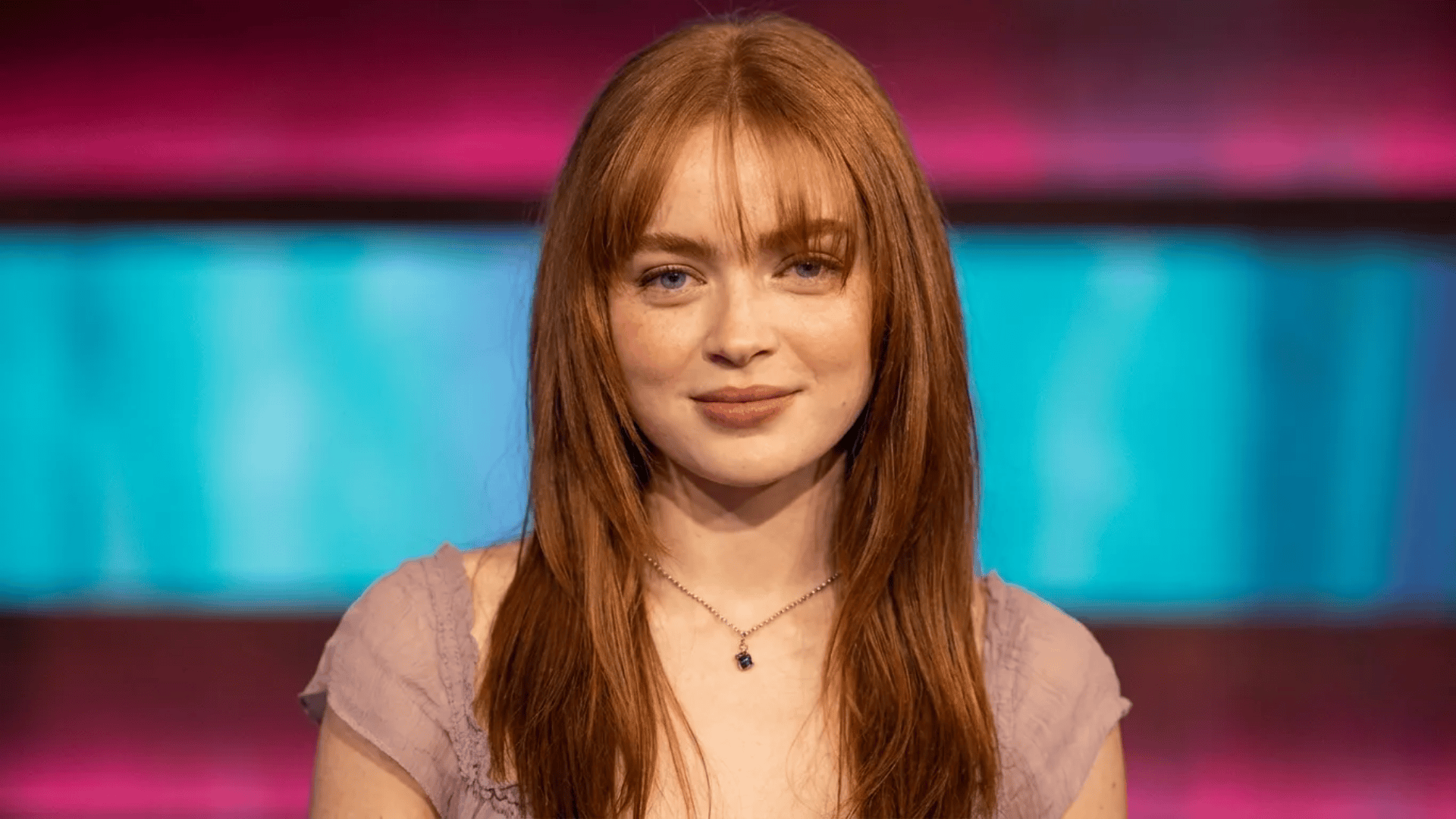 Woman with long straight red hair and light bangs, wearing a delicate necklace and a soft pink top, looking at the camera against a colorful blurred background.