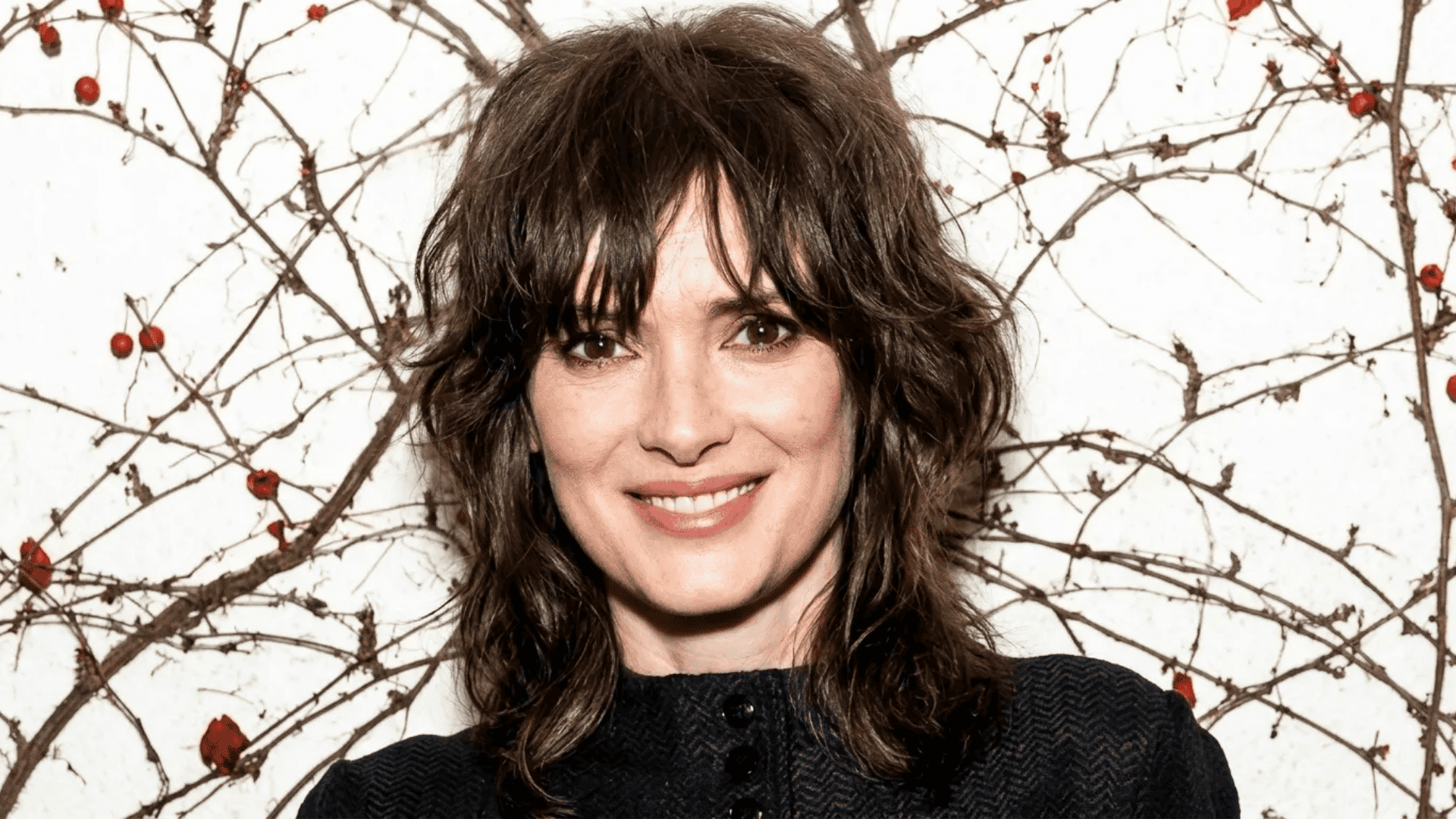 Woman with shaggy, layered dark brown hair and bangs, smiling at the camera, wearing a black top, with a decorative backdrop of branches and small red berries.