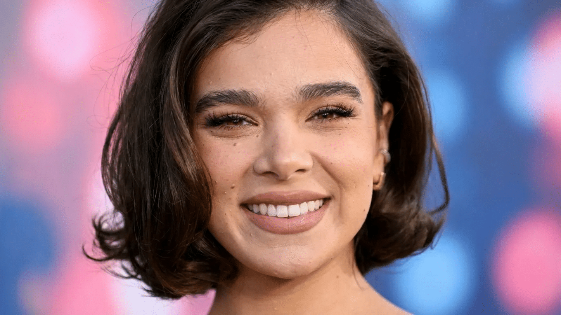 Woman with short, softly curled dark brown bob, smiling warmly, with natural makeup and small earrings, against a blurred pink and blue background.