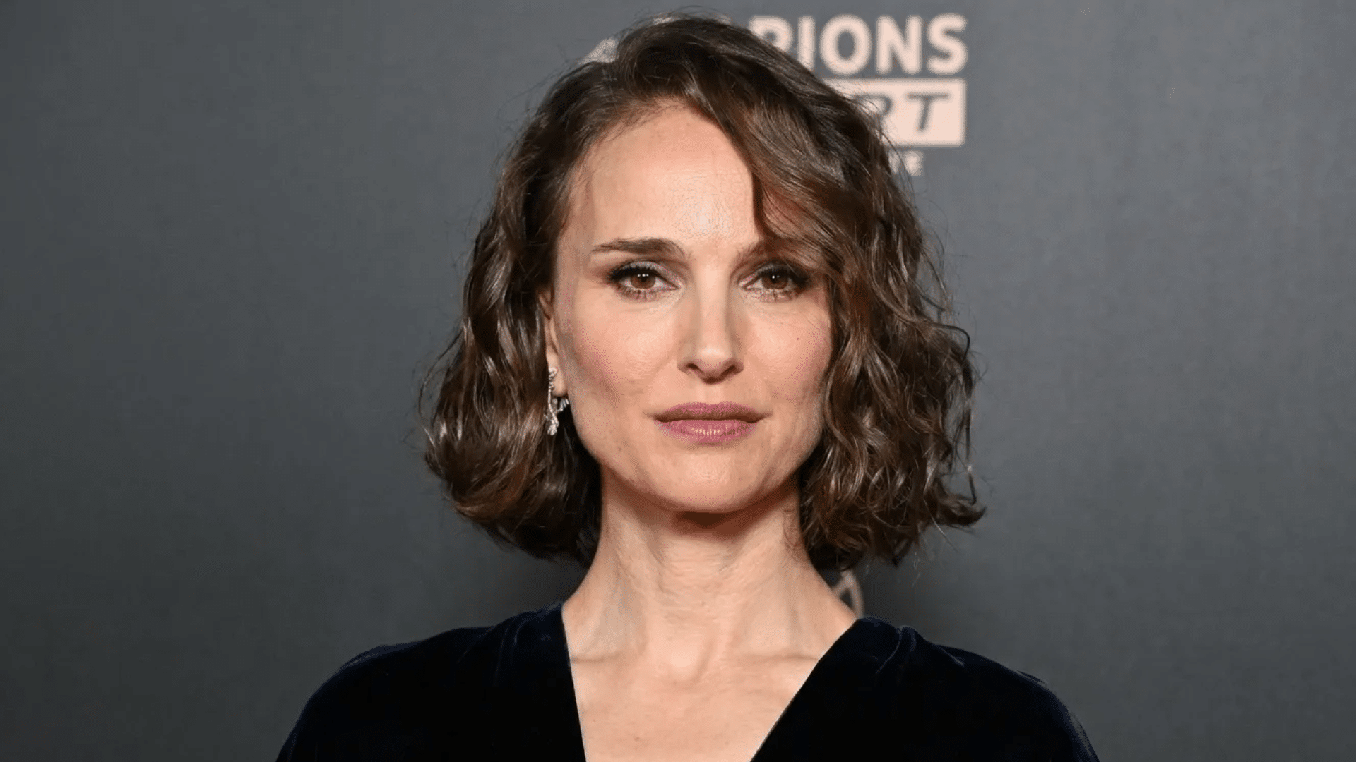 Woman with short, wavy brown bob haircut, wearing a dark outfit and subtle makeup, looking directly at the camera against a gray event-style backdrop.