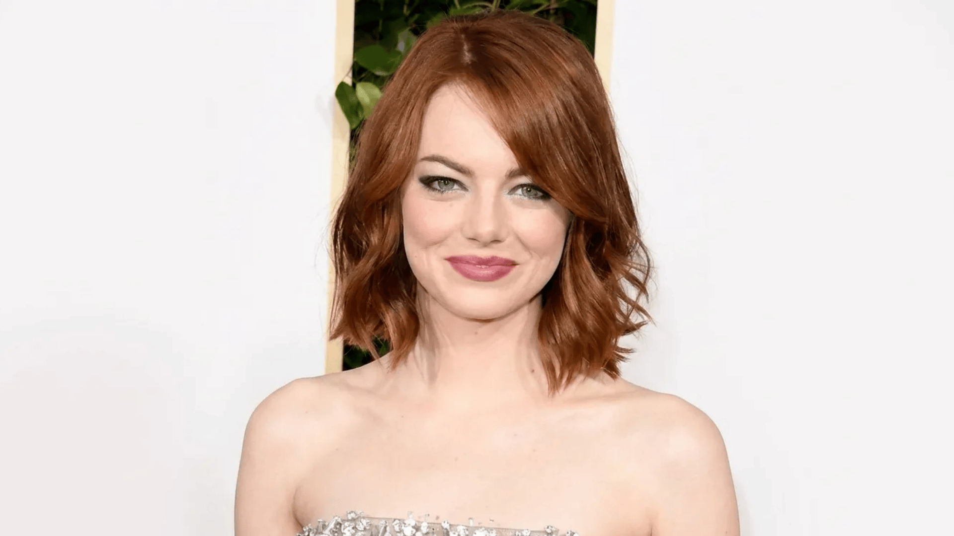 Woman with short, wavy copper-red bob haircut, wearing a strapless embellished dress, smiling at the camera against a light background with greenery.