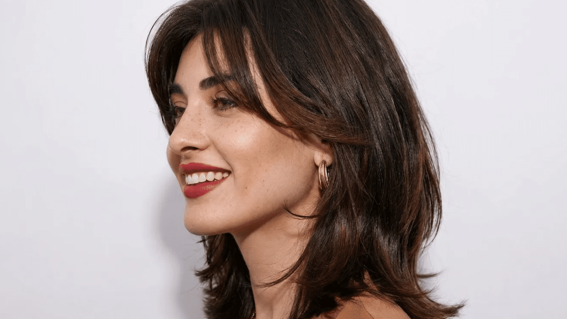 Woman with shoulder-length dark brown hair and soft bangs, smiling in profile, wearing gold hoop earrings and red lipstick against a neutral background.