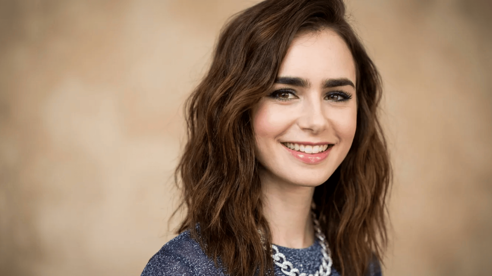 Woman with shoulder-length wavy brown hair, smiling, wearing a textured top and chain necklace, against a soft neutral background.