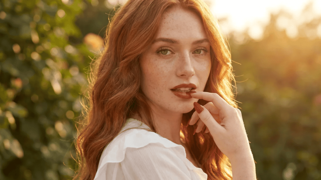 Woman with warm copper-red wavy hair, freckles, and soft makeup, posing outdoors in golden sunlight with a blurred green background.