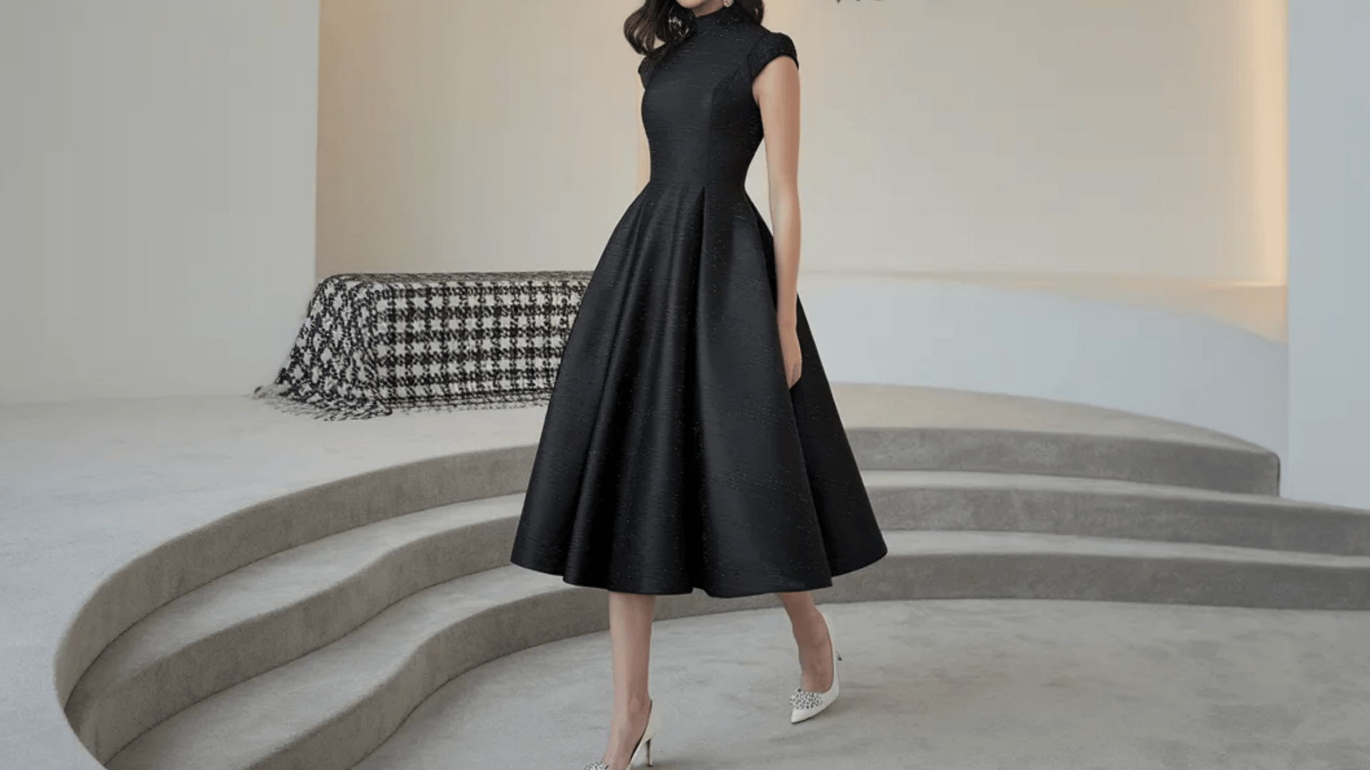 A woman wears a structured black midi dress with a high neck and flared skirt, paired with white pointed-toe heels.