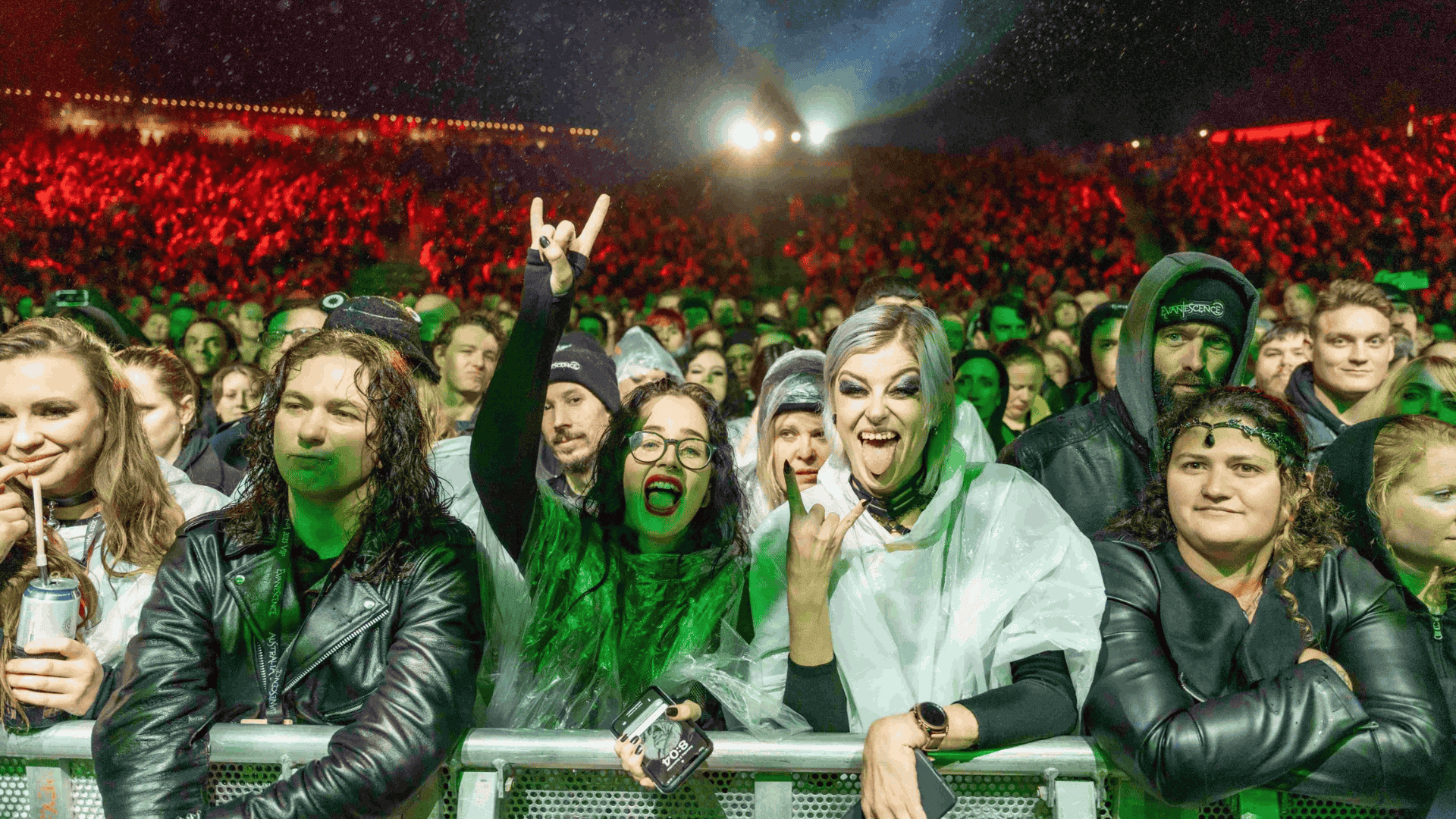 Crowd of concertgoers at a nighttime music festival cheering at the front barrier under colorful stage lights