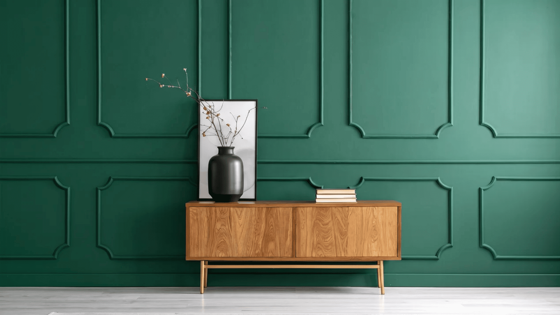 Deep green accent wall with decorative panel moulding, paired with a wooden sideboard, black vase with branches, framed art, and a small stack of books in a minimalist setting.