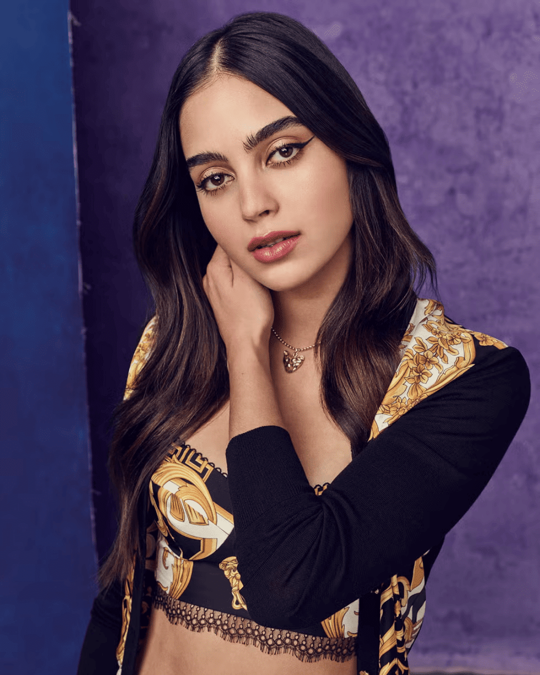 Portrait of a woman with long dark hair posing against a purple background, wearing a patterned outfit and necklace