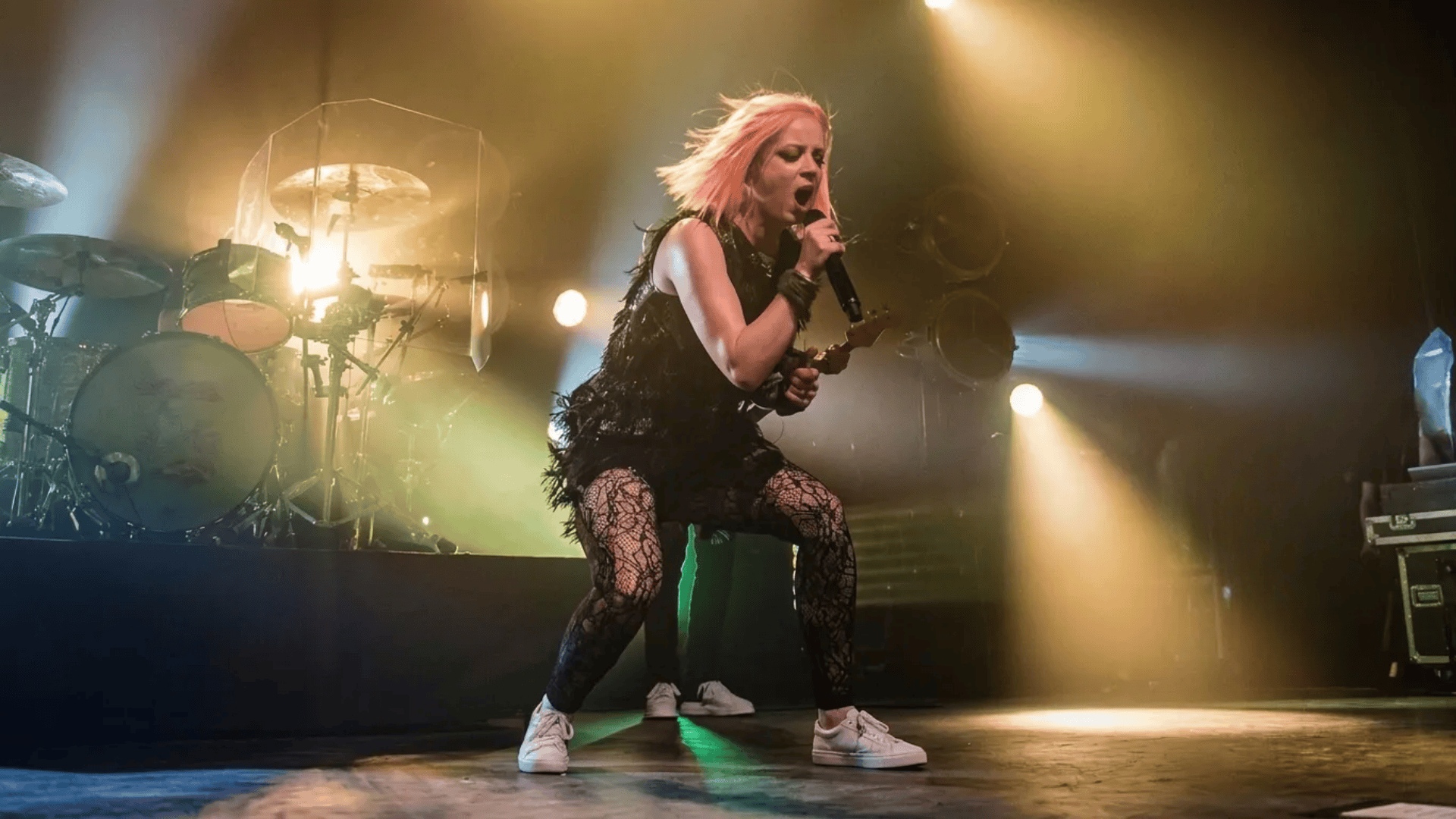 Singer Shirley Manson with pink hair performing on stage, holding a microphone, dramatic lighting, band setup visible in the background