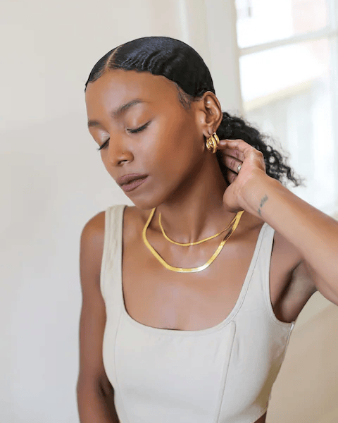 A woman in a beige top adjusts her gold earring while wearing matching jewelry.