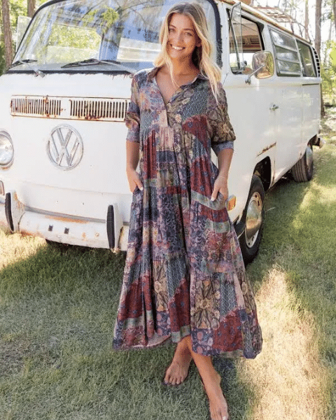 A woman in a patterned boho dress stands barefoot in front of a vintage van outdoors.