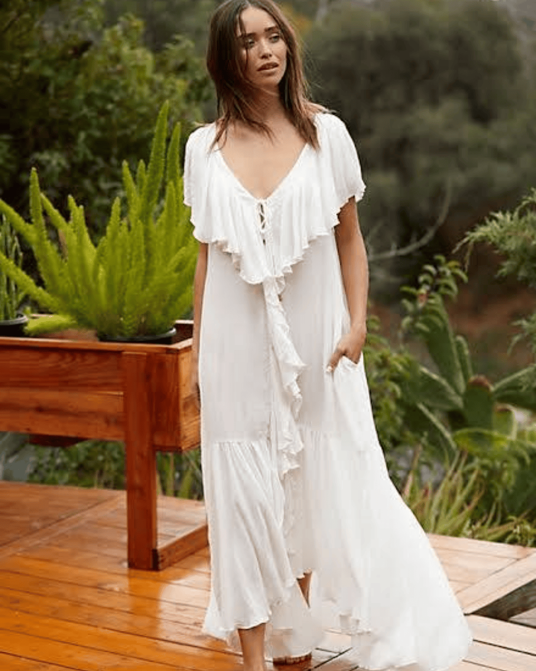 A woman standing barefoot on a wooden deck, wearing a flowy white dress with ruffled edges and a button-down front. She is surrounded by greenery and appears to be in a natural, outdoor setting.