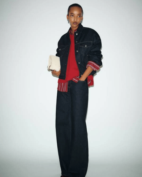 A woman wearing a red sweater layered over a striped shirt, paired with a dark denim jacket and wide-leg denim pants. She holds a small cream-colored handbag and stands against a plain white backg