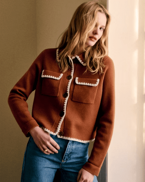 A woman wearing a rust-colored cardigan with white trim and button details, paired with light blue jeans. She stands by a window, with soft natural light highlighting her and the surroundings.