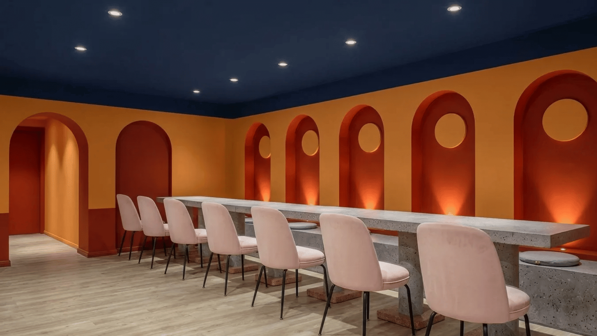 Bold modern interior with rhythmic arched wall details, warm ambient lighting, contrasting colors, and minimalist seating arranged around a long communal table.