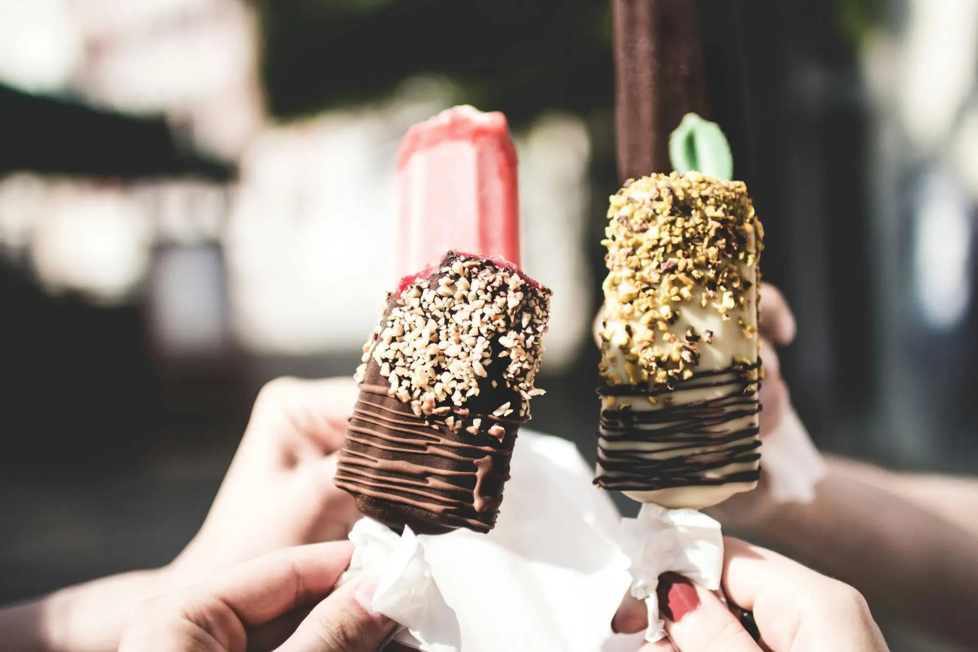 Three hands holding assorted ice cream bars with nuts and chocolate coating in sunny outdoor setting