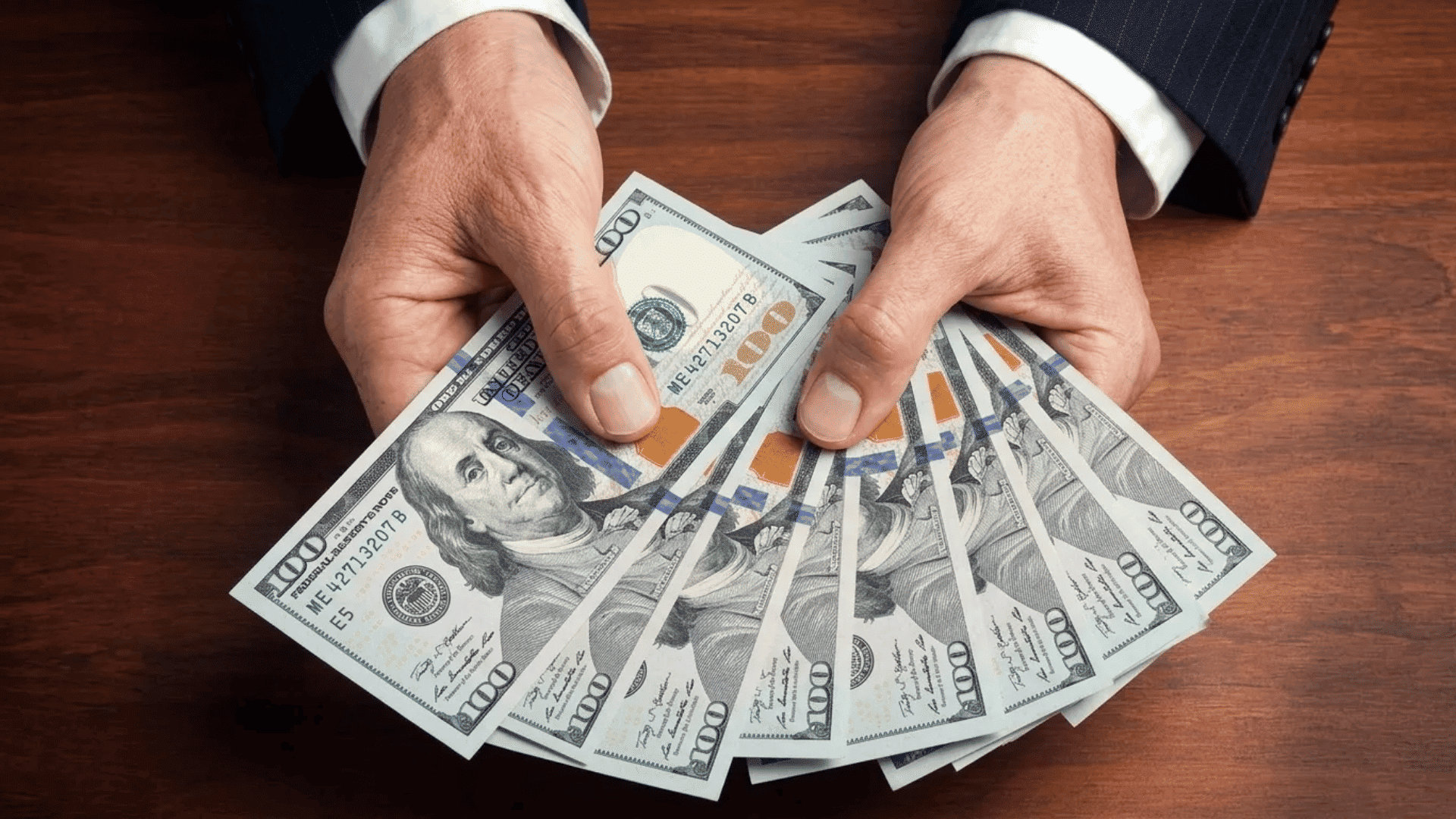 Hands in a business suit holding and fanning out several U.S. 0 bills over a wooden table.