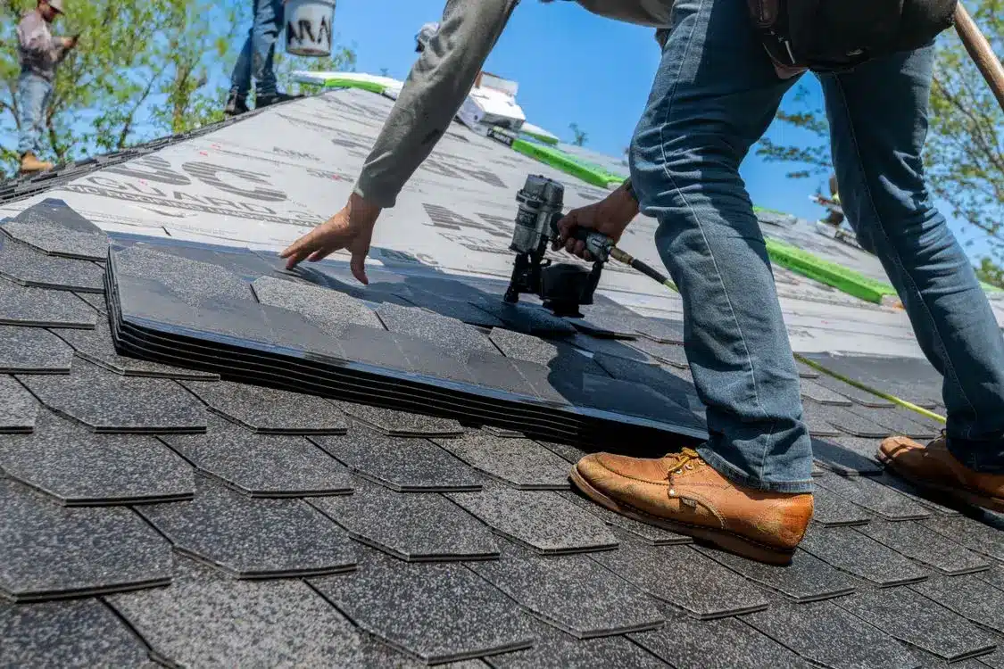 Worker installing asphalt shingles on a roof with a nail gun under clear blue sky