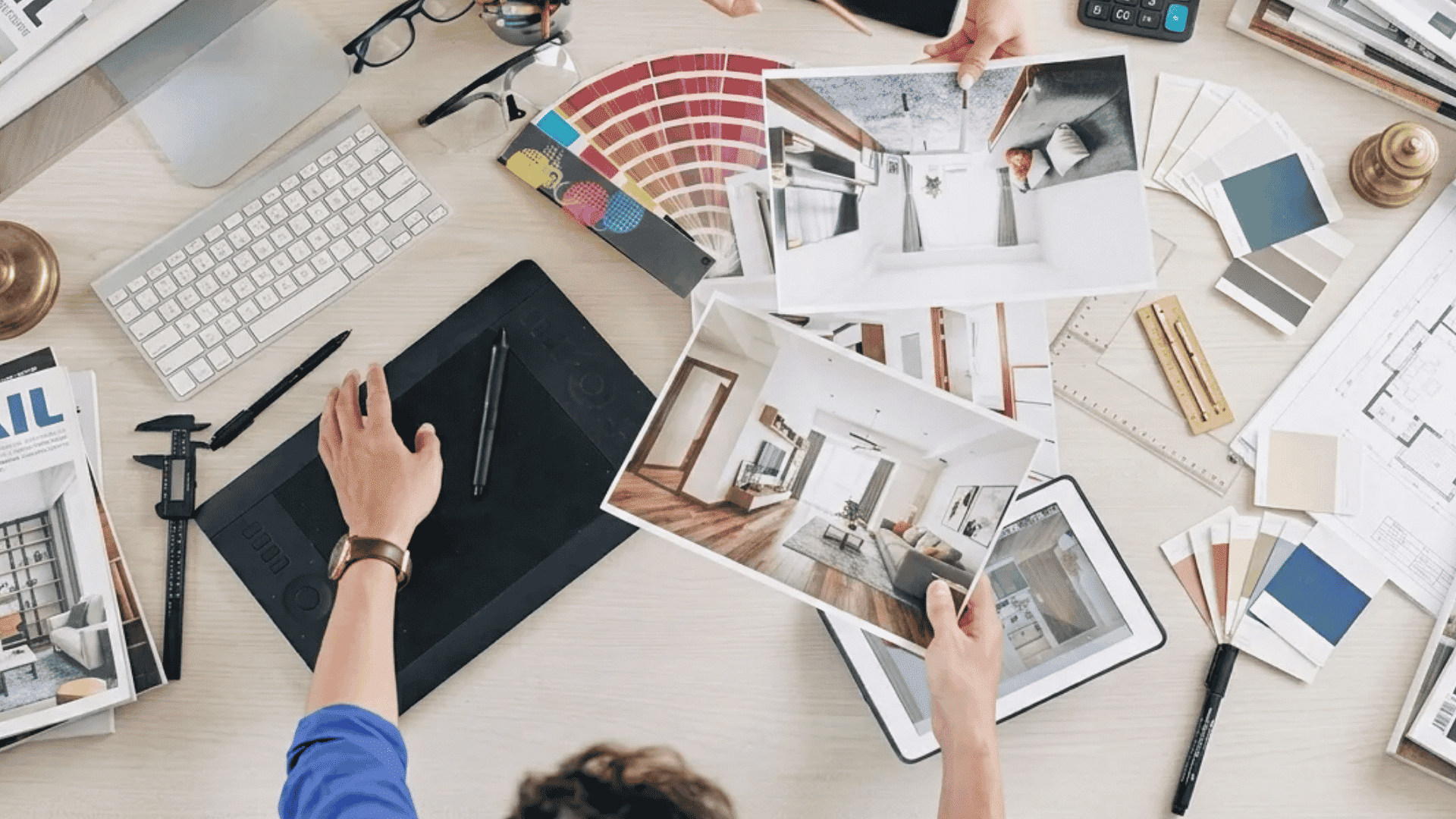 Top view of an interior design workspace with color swatches, room layout photos, a drawing tablet, keyboard, calculator, and design tools spread across a desk. (1)