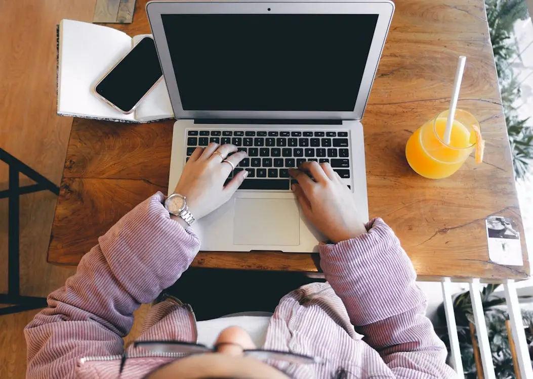 Person in striped sweater typing on laptop at wooden table with orange juice and smartphone
