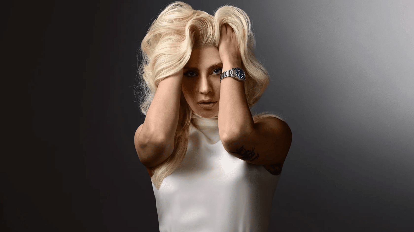 Lady Gaga in white top posing with hands in hair, wearing wristwatch, portrait against dark gradient background