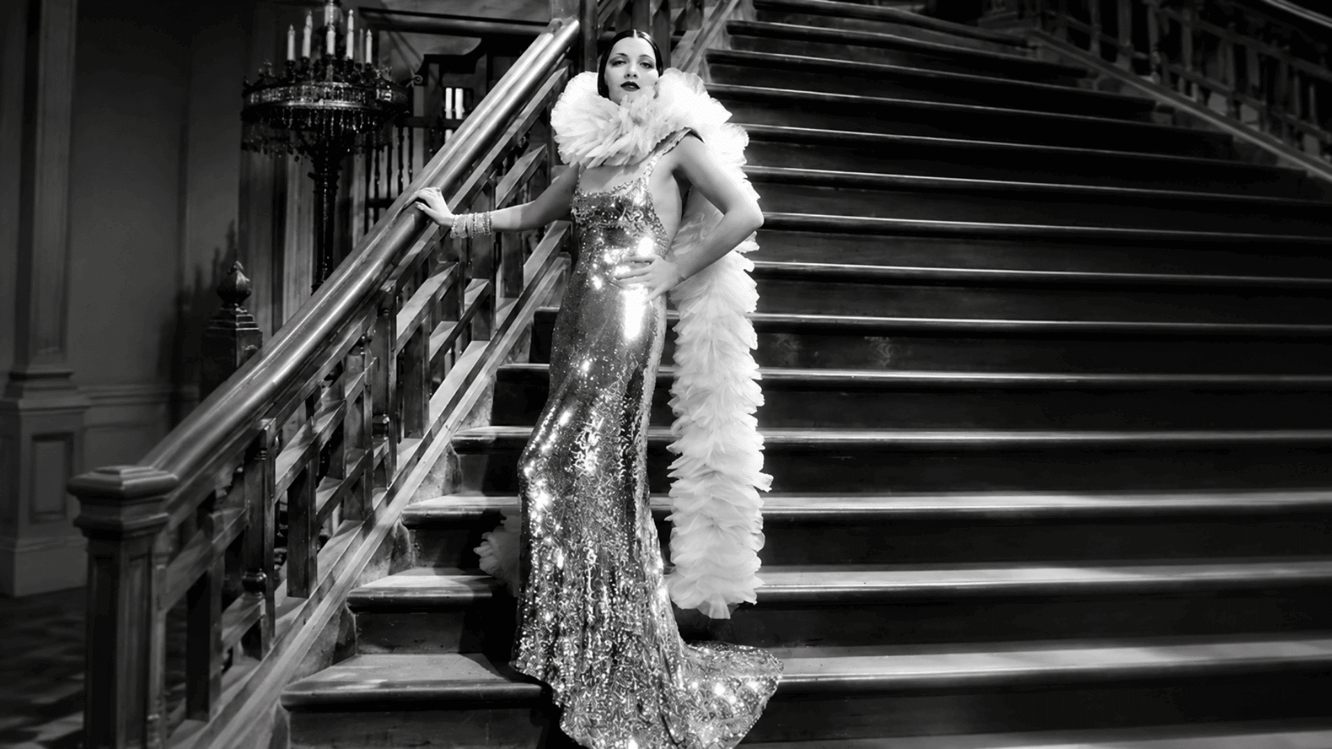 Black-and-white image of a woman in a sequined gown and feather boa standing on a grand staircase in a vintage Hollywood style pose.