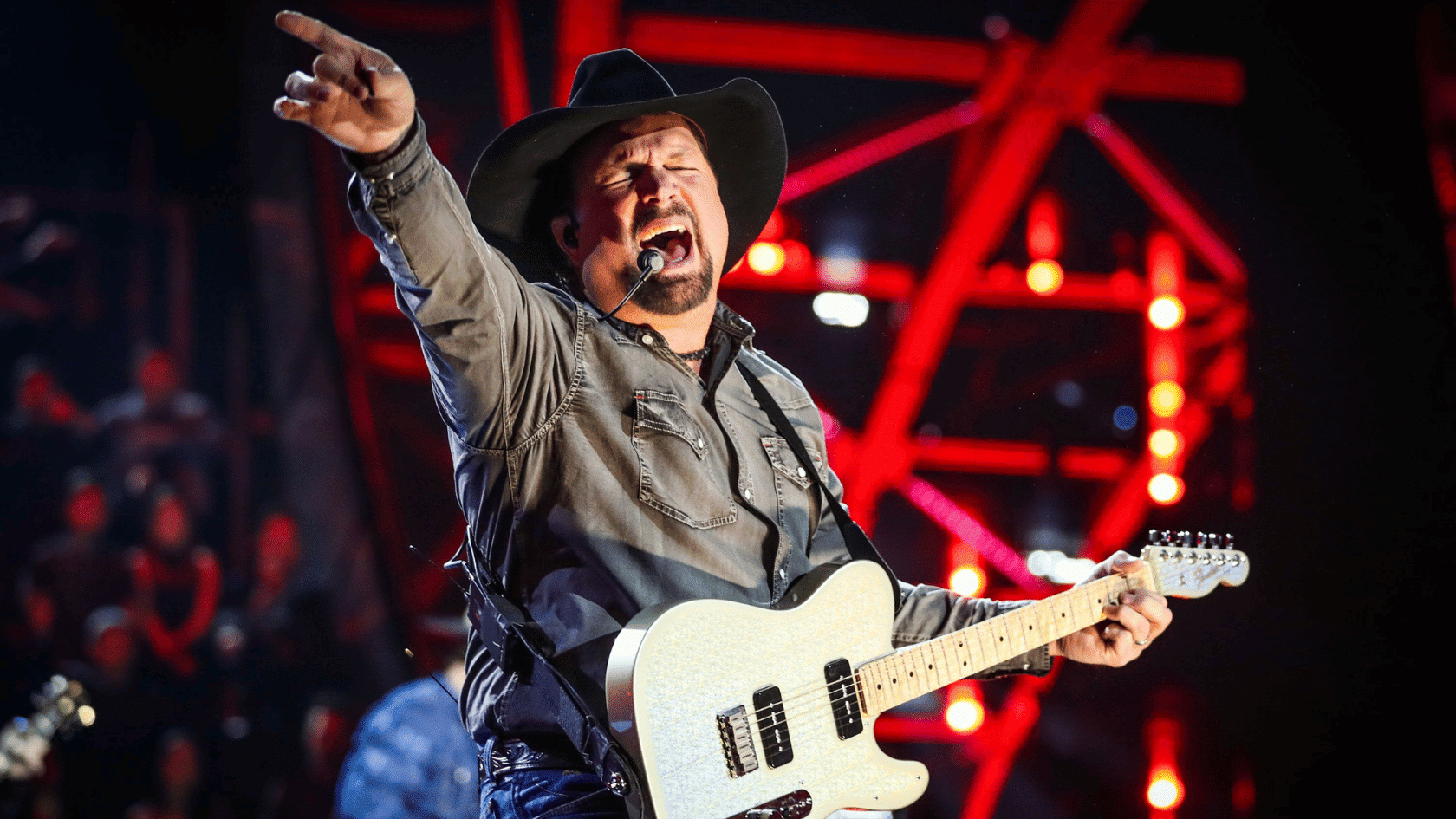 Garth Brooks performing on stage, singing into a headset mic while playing electric guitar and pointing to crowd