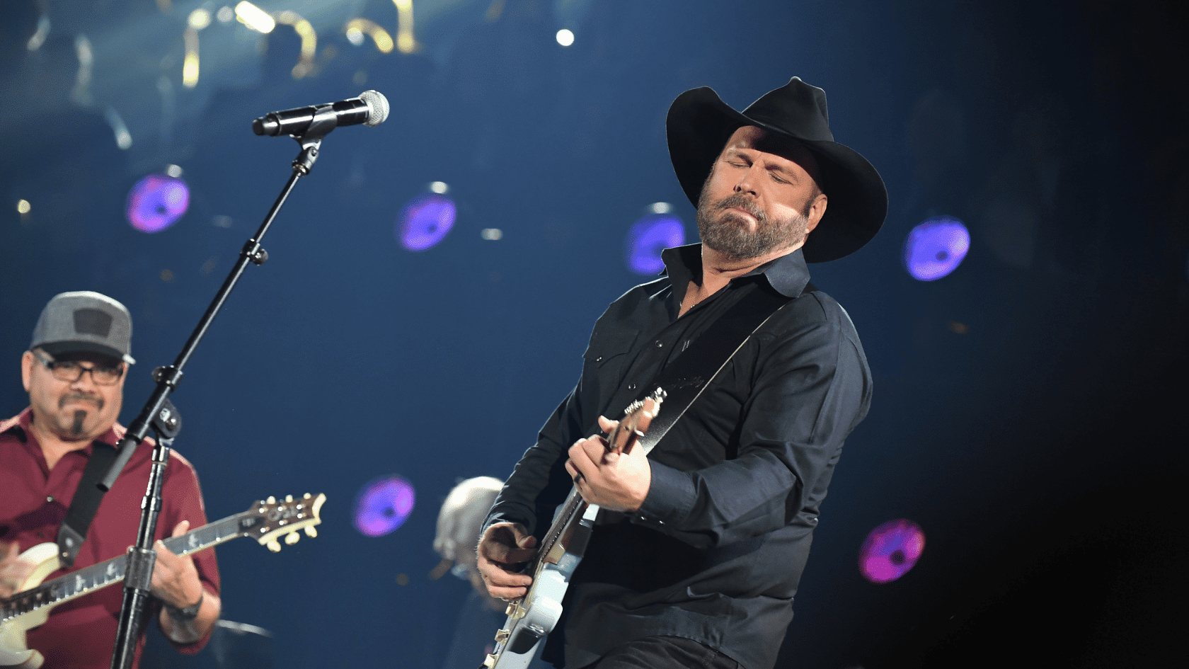 Garth Brooks performing live on stage, playing electric guitar with eyes closed under dramatic concert lighting.