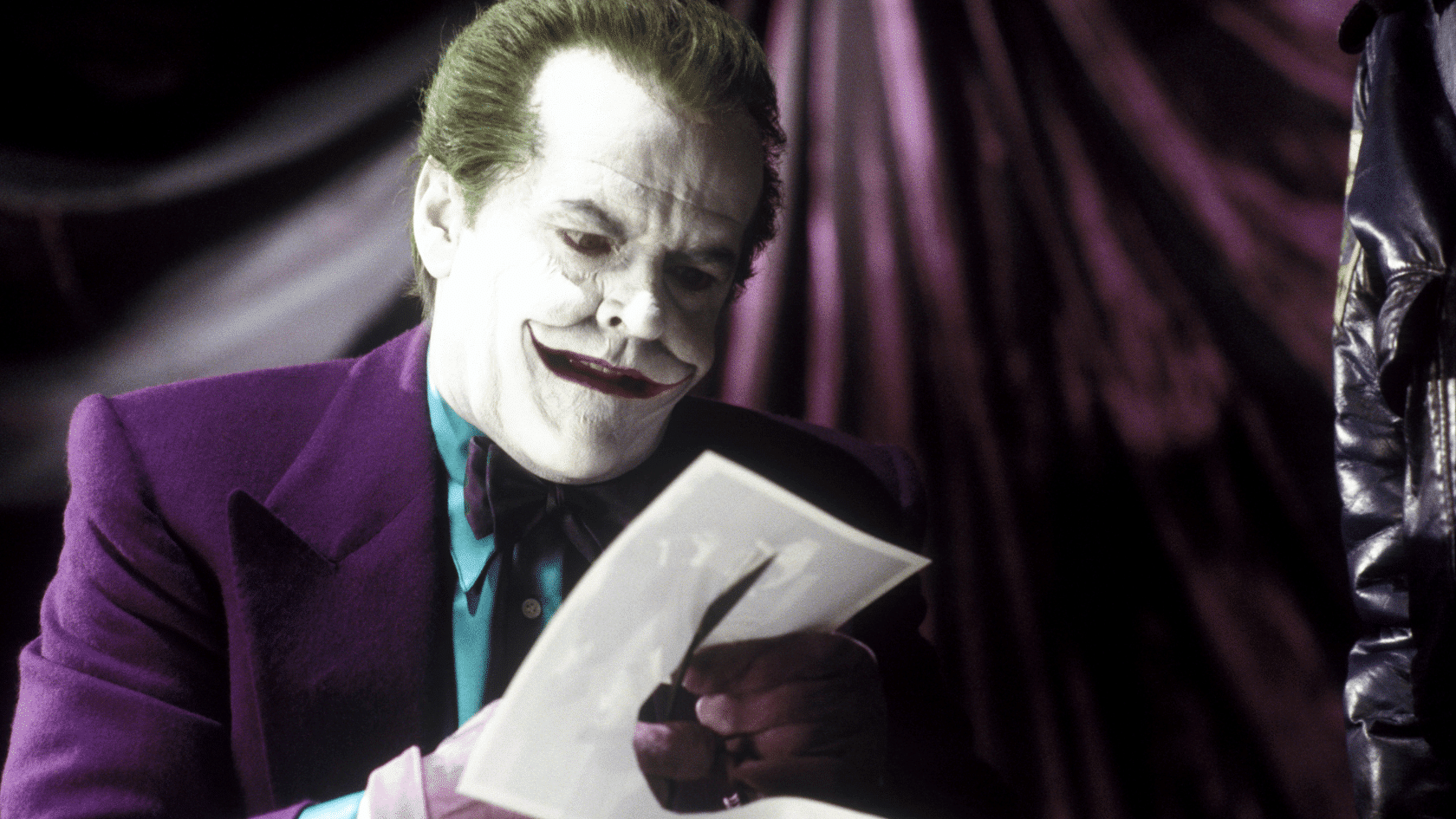Jack Nicholson as the Joker in a purple suit reading a letter with a sinister smile against a dark draped background