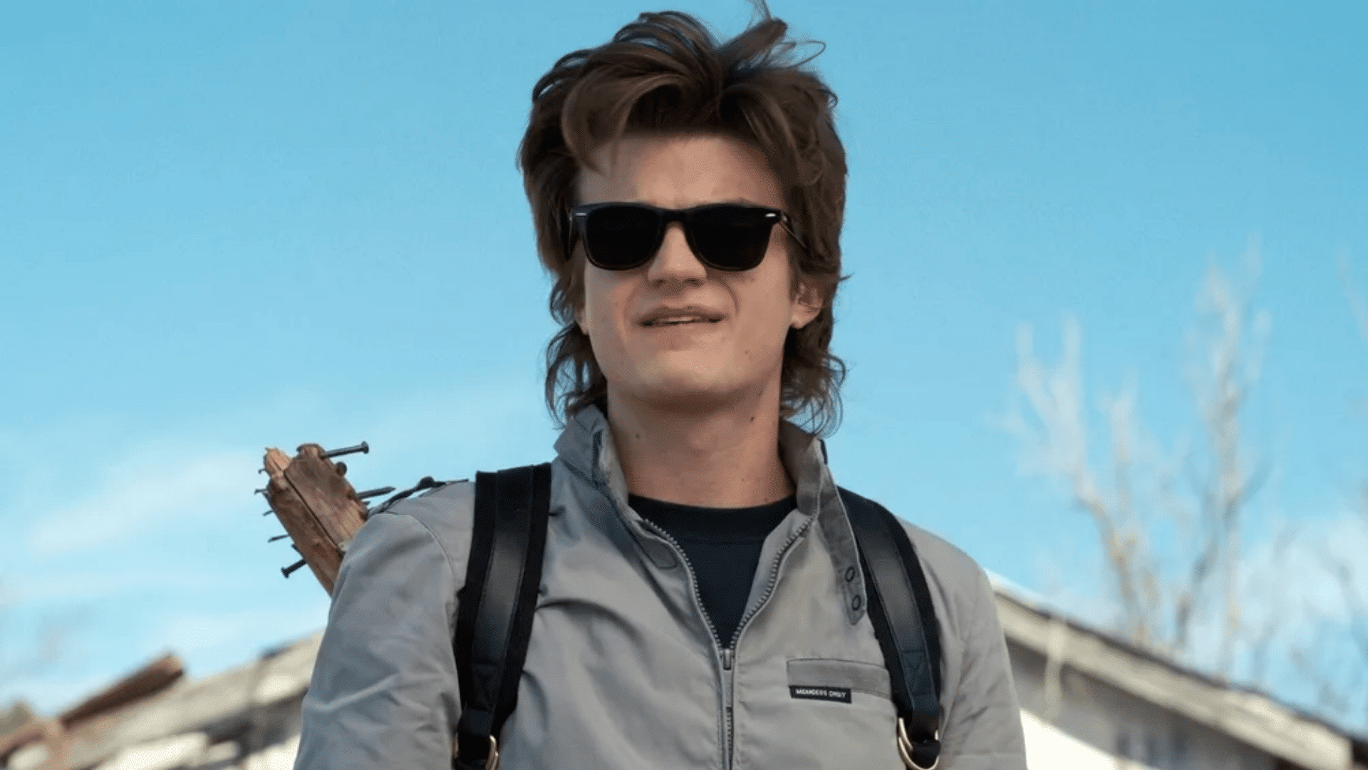 Joe Keery as Steve Harrington in Stranger Things wearing sunglasses with short mullet haircut, textured layers and longer back, standing outdoors with backpack.