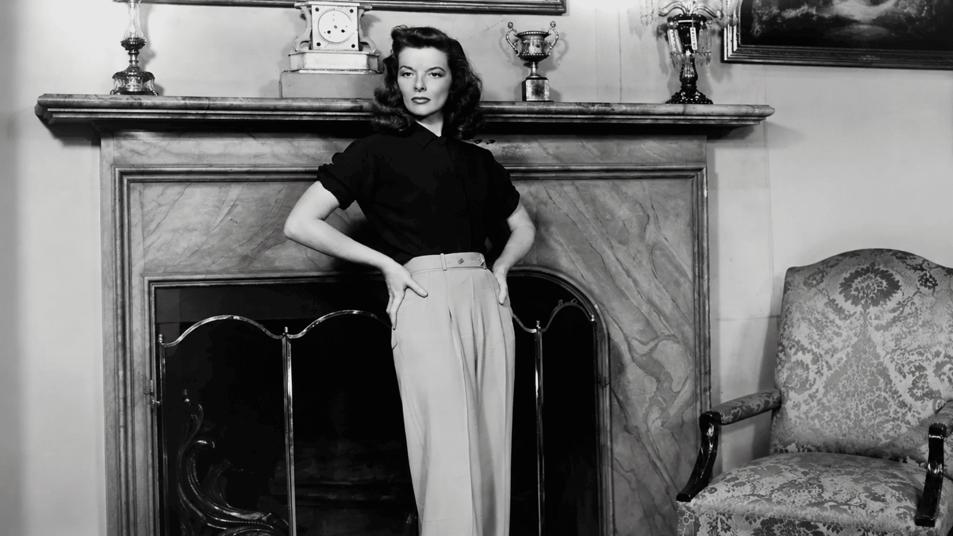 Black-and-white portrait of a woman in a blouse and high-waisted trousers standing in a vintage interior.