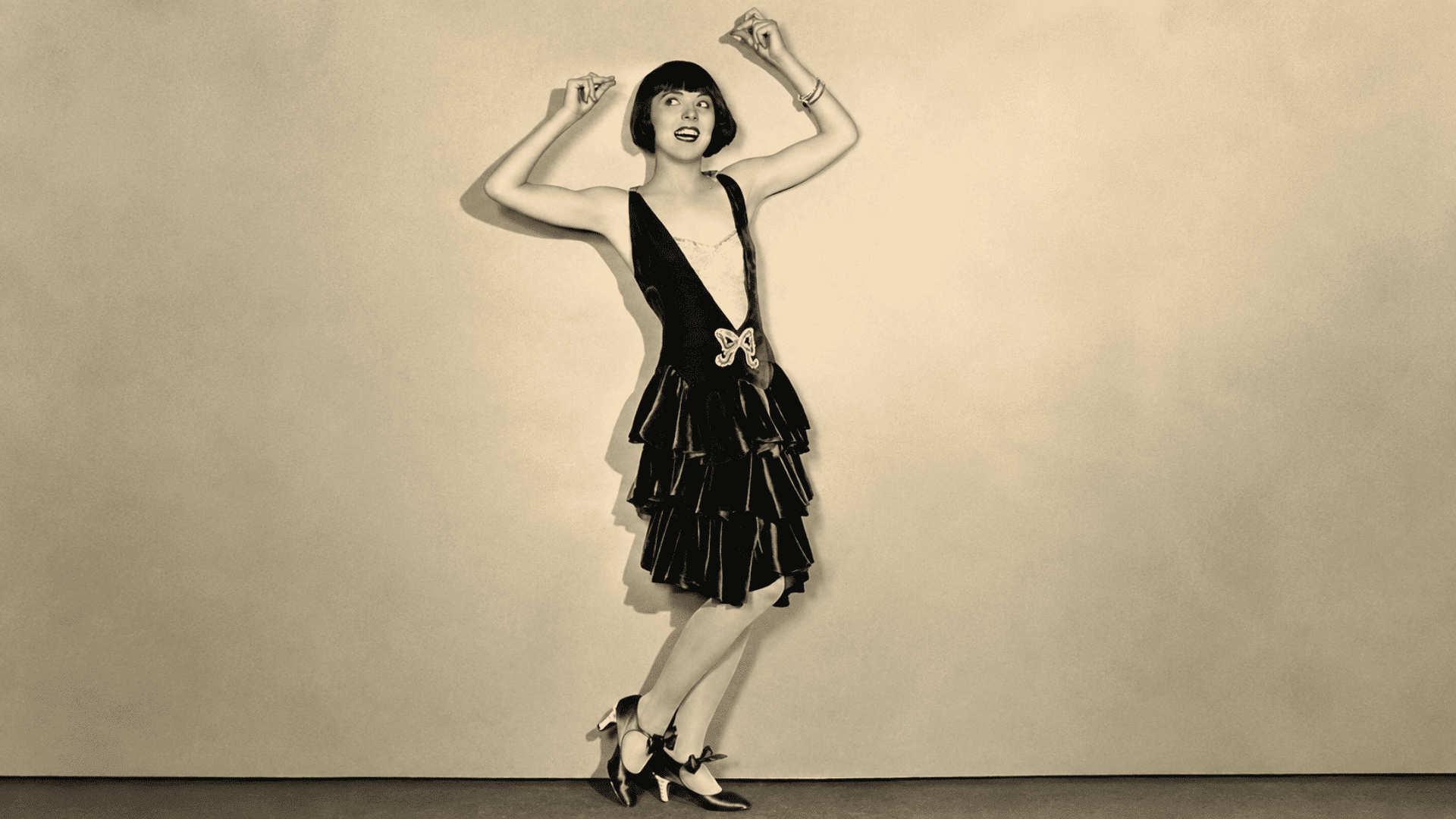 Young woman in a 1920s flapper dress posing mid-dance with raised arms in a sepia-toned studio portrait.