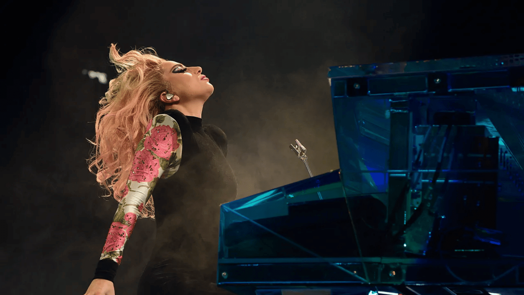 Lady Gaga performing on stage with pink hair, leaning back while playing a blue piano under dramatic stage lighting and smoke.
