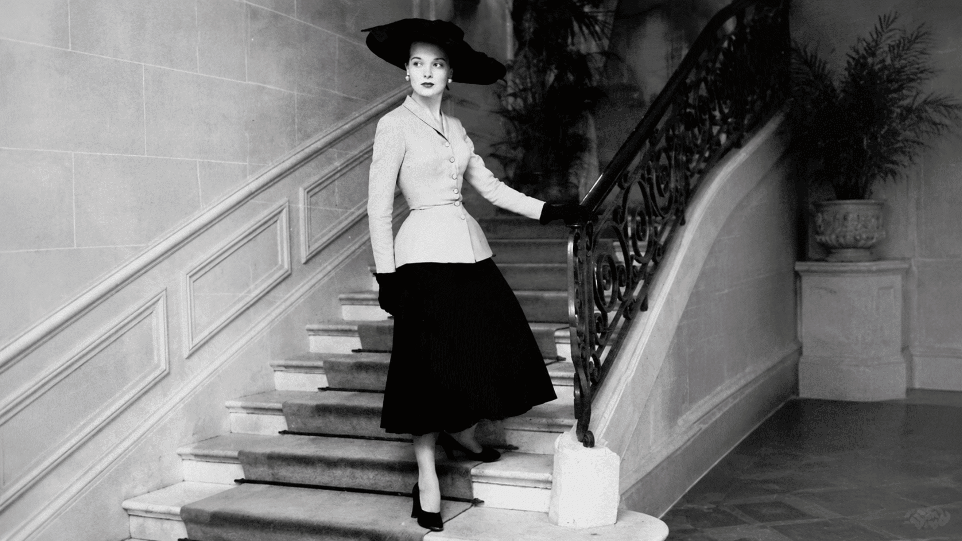 Woman in vintage outfit standing on a staircase, holding the railing, wearing a wide-brim hat and tailored jacket in a black-and-white portrait.