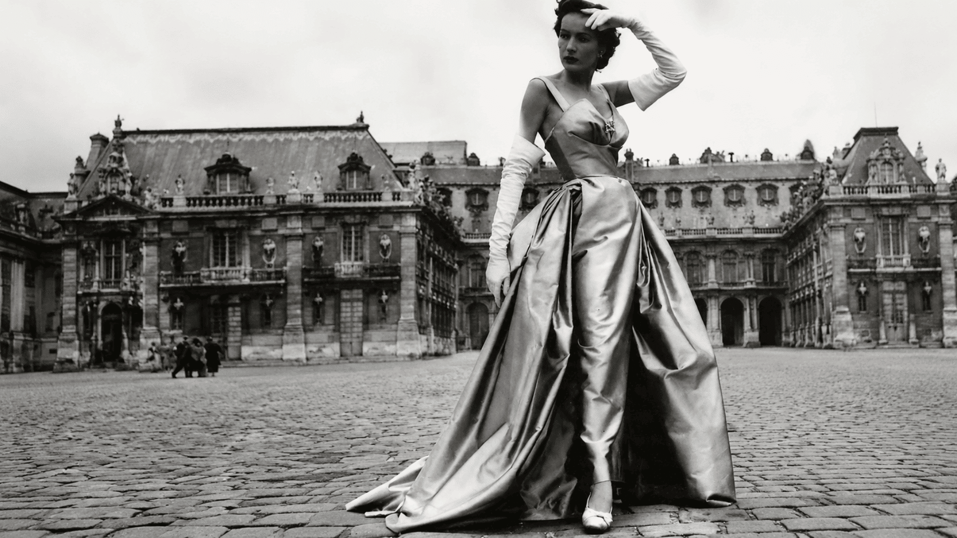 Woman in satin gown posing in a grand cobblestone plaza with historic buildings in a vintage black-and-white fashion photograph.