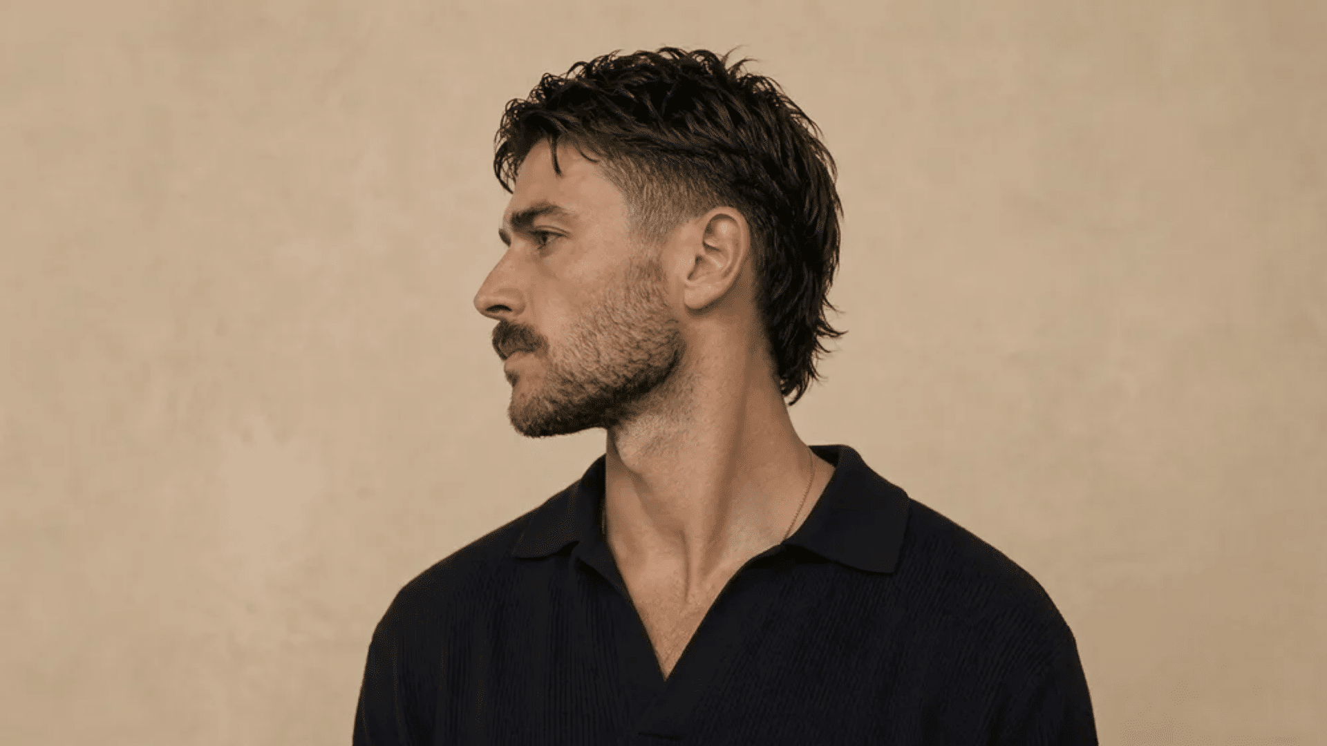 Man in side profile with short mullet haircut, featuring faded sides, textured top, and slightly longer back against a beige background.