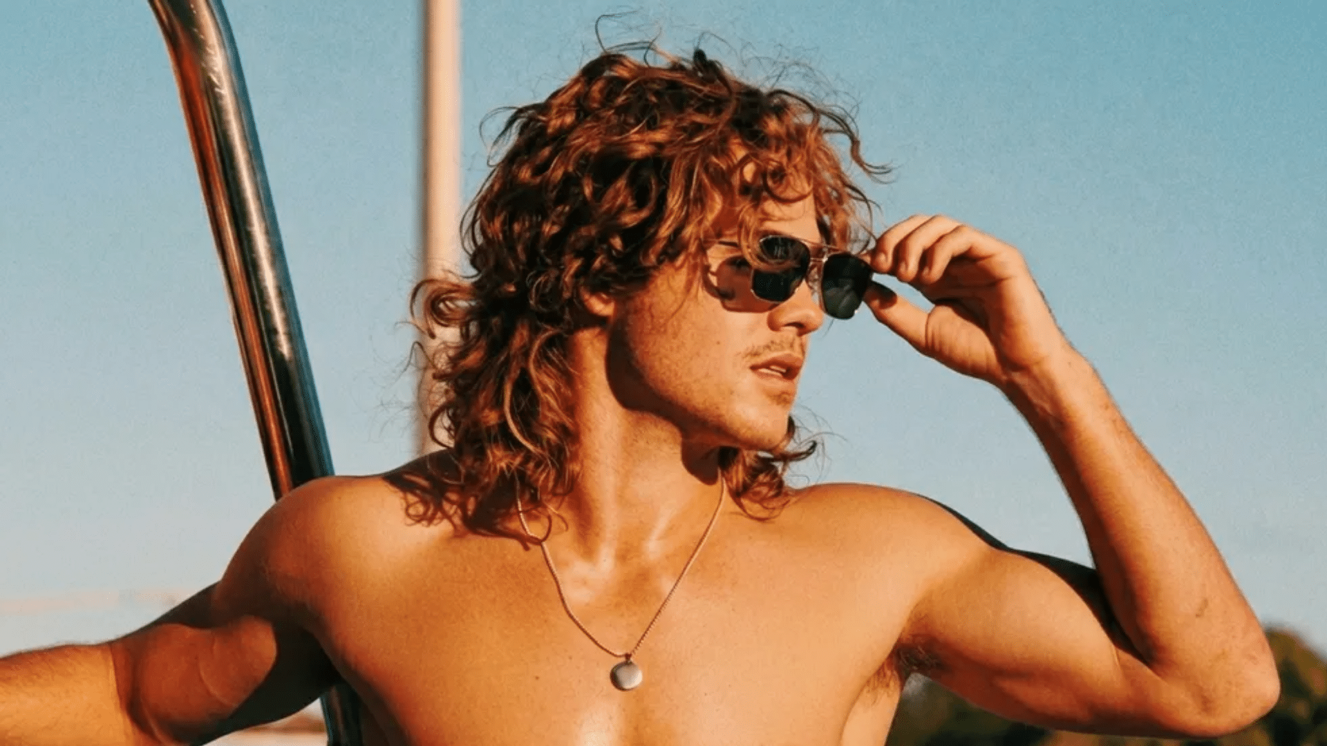 Dacre Montgomery as Billy Hargrove with curly mullet hairstyle wearing sunglasses, adjusting them while standing outdoors in sunlight with a relaxed pose.