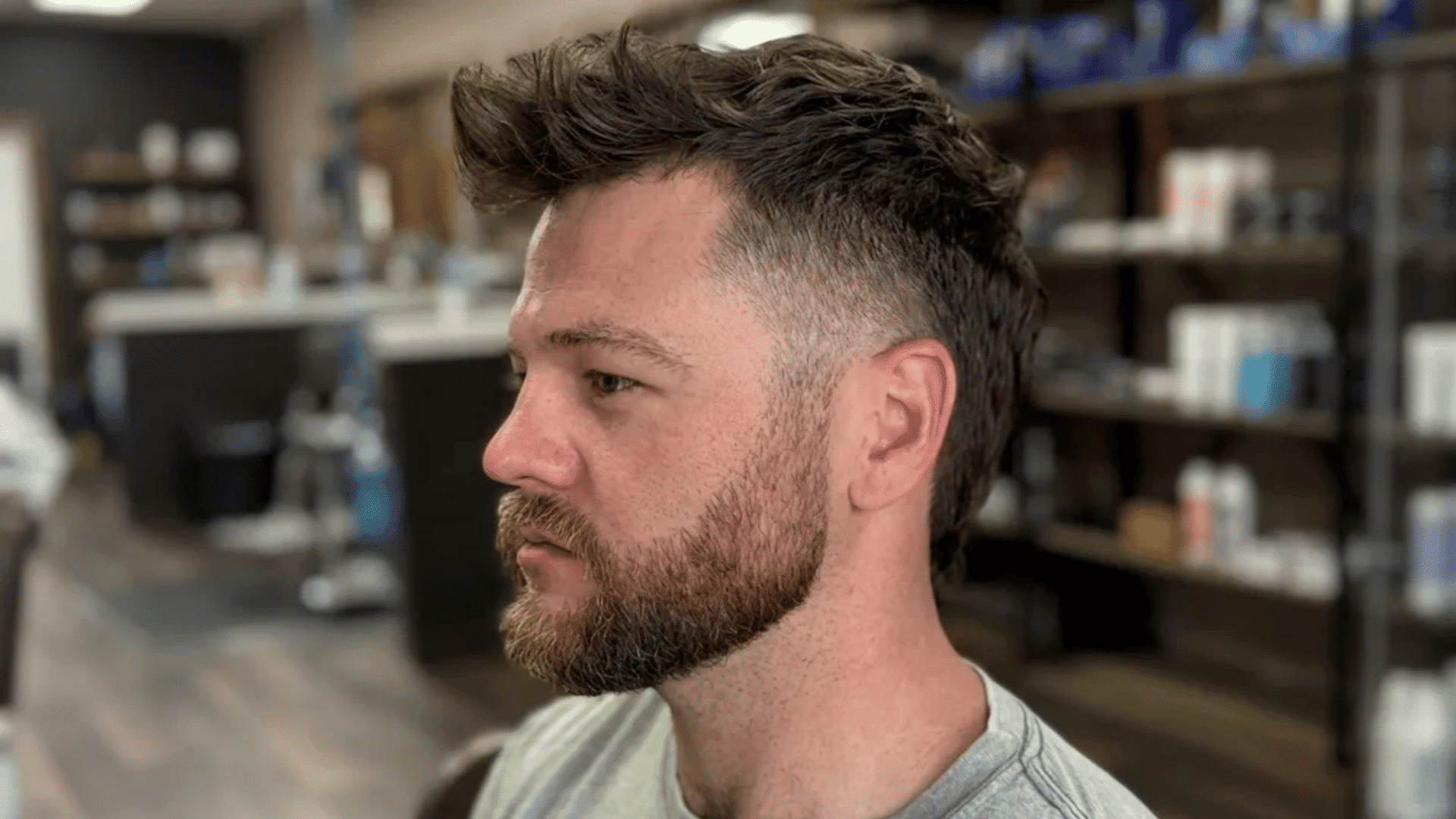 Man with short mullet haircut, textured top with faded sides and fuller beard, shown in profile inside a barbershop.