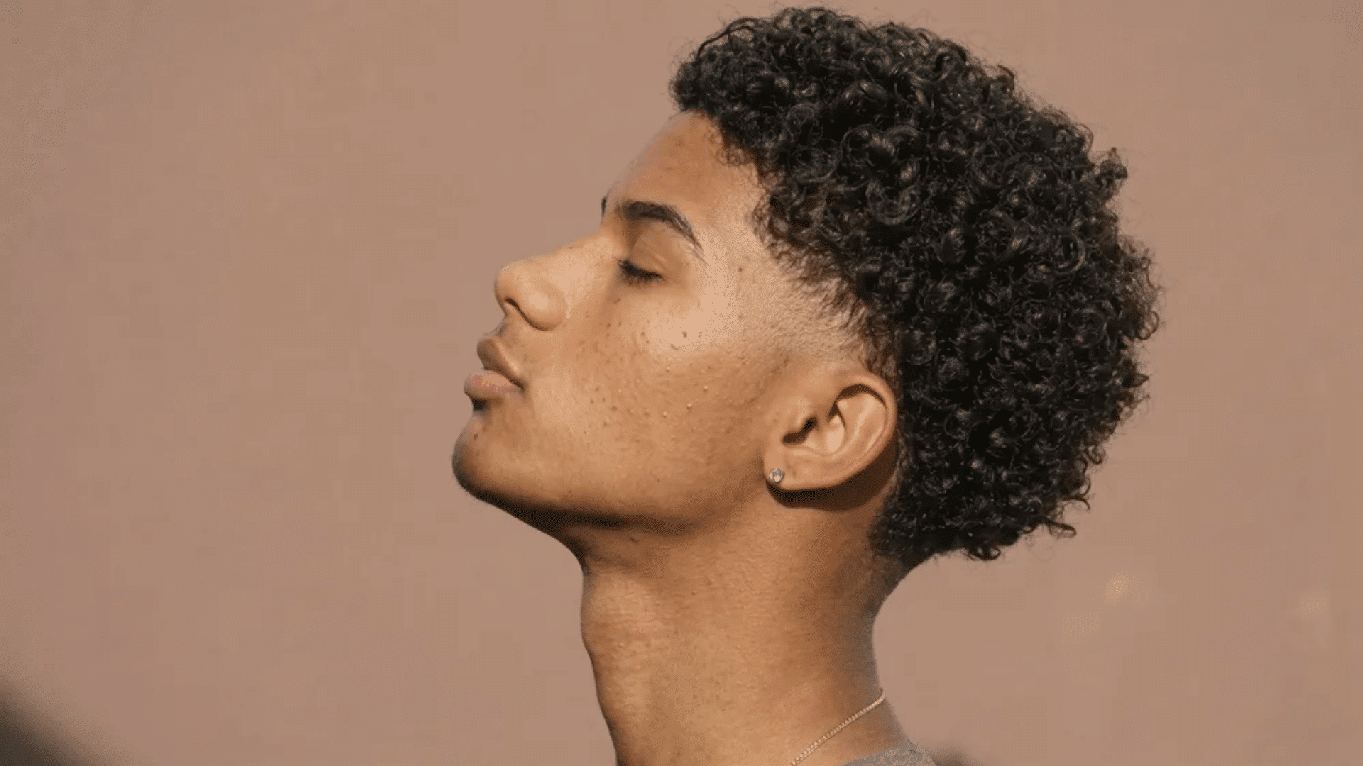 mixed boys haircuts curly top with high fade side profile of young man with tight curls against plain neutral background