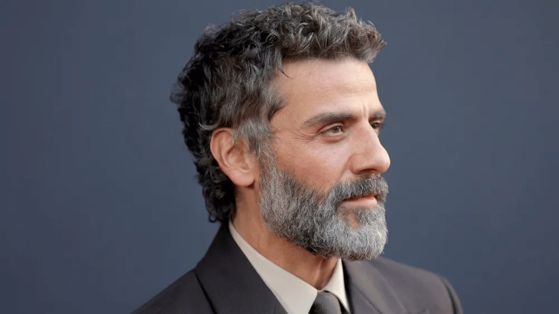 Oscar Issac with short mullet haircut, gray beard, wearing suit against dark blue background.