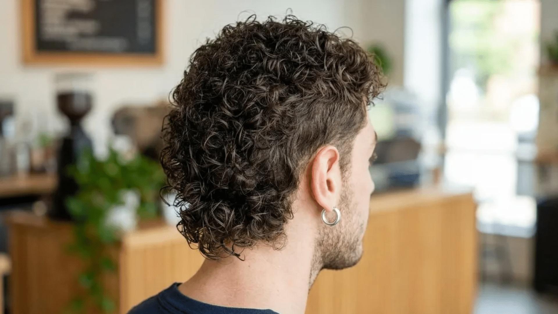 Short mullet haircut on curly hair, back view showing tight curls, faded sides, and longer textured back section inside a barbershop.