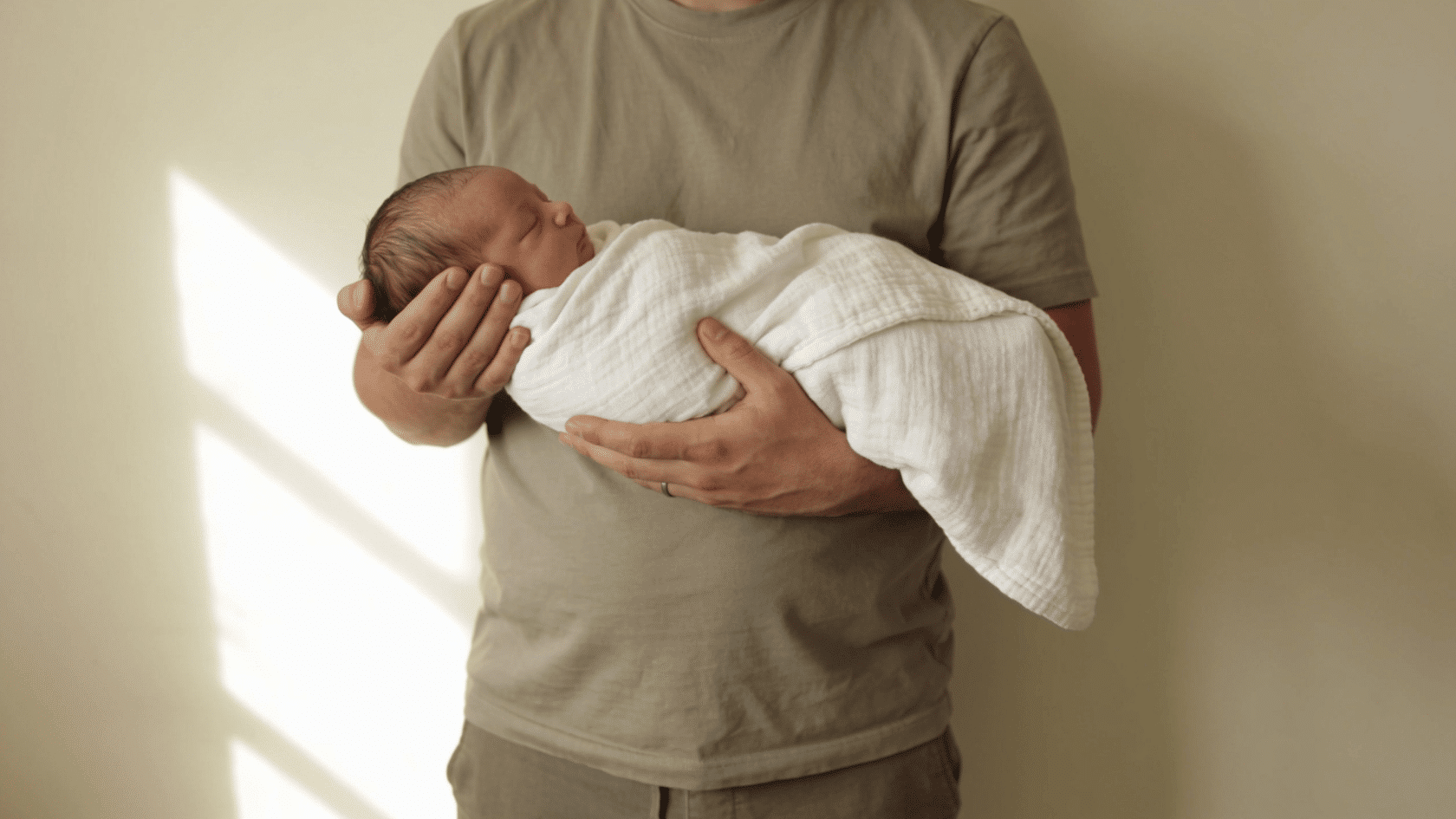 Father wearing a plain neutral shirt and pants in a simple indoor newborn photoshoot setting