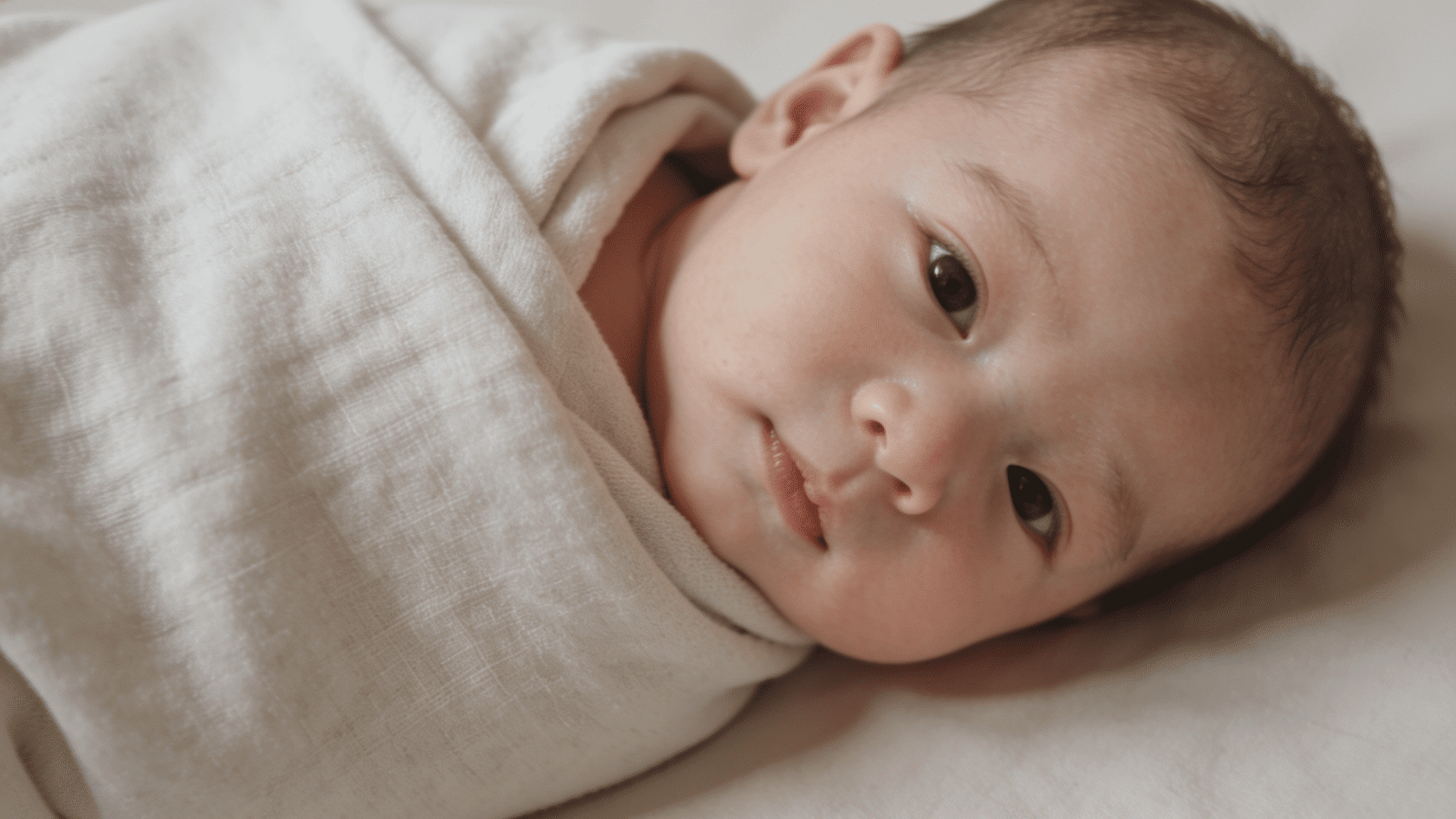 Baby wrapped in a plain swaddle blanket lying in a neutral indoor setting