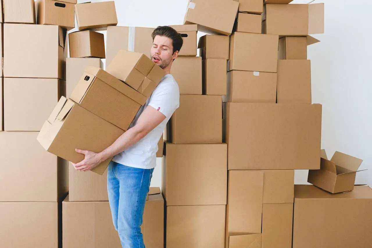 Man carrying multiple cardboard boxes in front of a large stack indoors