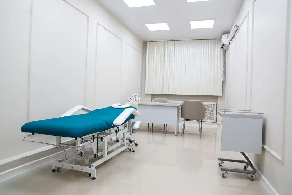 Medical examination room with teal examination table, desk, and chair under fluorescent lighting