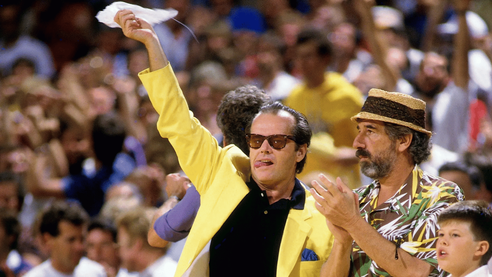 Jack Nicholson at a basketball game in a yellow jacket, wearing sunglasses and waving a towel in the crowd